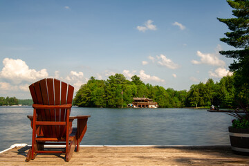 Wall Mural - On the wooden dock, a lone Muskoka chair offers a tranquil view of the serene lake. Across the water, a cottage is nestled amidst a lush backdrop of green trees.