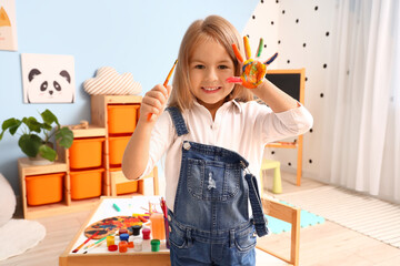 Wall Mural - Cute little girl with her hand in paint and brush at home