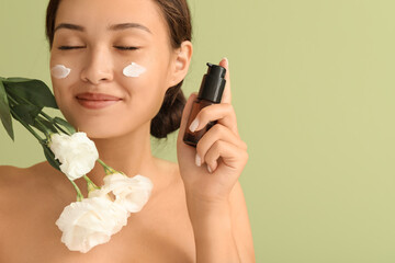 Wall Mural - Beautiful Asian woman with facial cream and flowers on green background, closeup