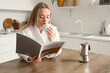 Pretty young woman reading magazine while drinking coffee in modern kitchen