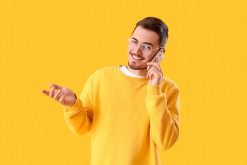 Wall Mural - Young man in eyeglasses talking by mobile phone on yellow background