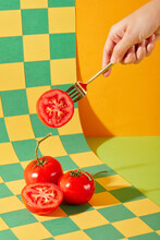 The Hand Is Holding A Metal Fork Pinned To Half Of A Tomato. Juicy Red Tomatoes Are Displayed On A Checkered Background With Two Main Colors: Green And Yellow. Art Space With Fresh Vegetables.