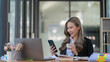 © Songsak C - An Asian businesswoman using a smart phone and holding a cup of coffee while sitting in her office room.