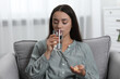 © New Africa - Depressed woman with glass of water taking antidepressant pills in armchair indoors