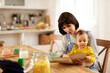 © Stockphotodirectors - Young mother reading a book to her baby boy while having having breakfast in the dining room