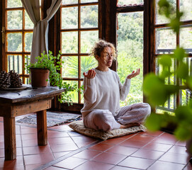 Full length attentive young Caucasian woman making mudra gesture, sitting in lotus position sitting on soft cushion on floor at home. Peaceful woman meditating deeply, doing breathing yoga 