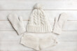 A white knitted hat with a pompom, woolen mittens and socks lie on a white wooden background. Details of winter clothing