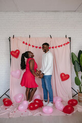 Wall Mural - Young African couple holding hands while standing on pink background with heart shaped balloons