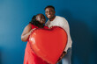 © gstockstudio - Happy young African couple holding red heart shaped balloon while bonding on blue background