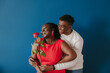 © gstockstudio - Happy young African man embracing his girlfriend holding red rose on blue background