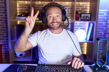 Poster - Middle age man with beard playing video games wearing headphones showing and pointing up with fingers number two while smiling confident and happy.