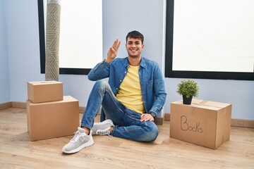 Wall Mural - Young hispanic man sitting on the floor at new home showing and pointing up with fingers number three while smiling confident and happy.