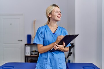 Poster - Young blonde woman pysiotherapist smiling confident writing on document at rehab clinic