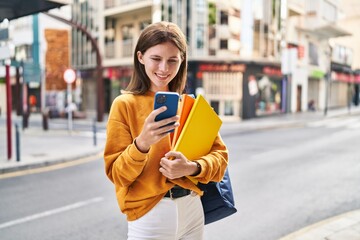 Sticker - Young blonde woman student using smartphone holding books at street