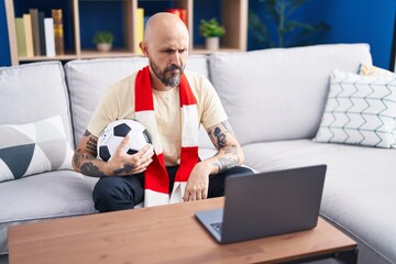 Sticker - Hispanic man with tattoos watching football match hooligan holding ball on the laptop thinking attitude and sober expression looking self confident