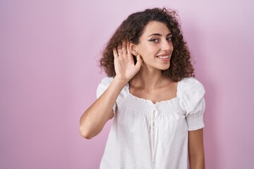 Sticker - Hispanic woman with curly hair standing over pink background smiling with hand over ear listening an hearing to rumor or gossip. deafness concept.