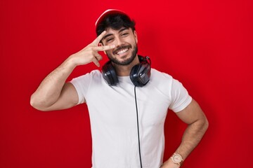 Poster - Hispanic man with beard wearing gamer hat and headphones doing peace symbol with fingers over face, smiling cheerful showing victory