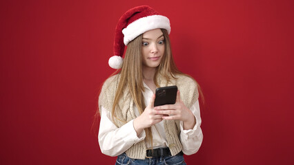 Poster - Young blonde woman wearing christmas hat using smartphone over isolated red background