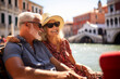 A middle-aged husband and wife sit in a gondola and admire the canals of Venice.