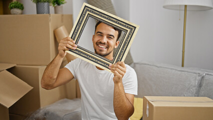 Young hispanic man holding vintage frame sitting on sofa smiling at new home