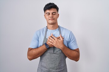 Sticker - Hispanic young man wearing apron over white background smiling with hands on chest with closed eyes and grateful gesture on face. health concept.