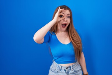 Sticker - Redhead woman standing over blue background doing ok gesture shocked with surprised face, eye looking through fingers. unbelieving expression.