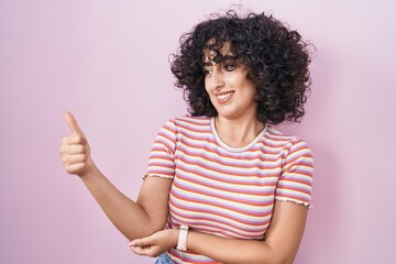 Wall Mural - Young middle east woman standing over pink background looking proud, smiling doing thumbs up gesture to the side