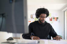 Black Man Working In The Office. Sitting At His Desk, He Is Checking Business Documents. He Wears A Casual Look, Has Afro Hair And A Beard