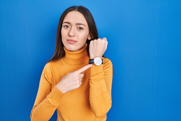 Sticker - Young brunette woman standing over blue background in hurry pointing to watch time, impatience, looking at the camera with relaxed expression