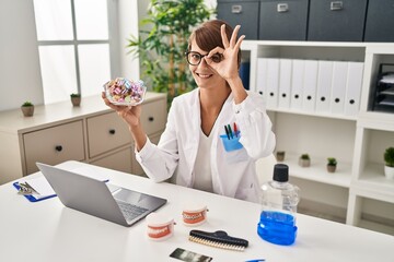 Wall Mural - Brunette dentist woman holding sweets smiling happy doing ok sign with hand on eye looking through fingers