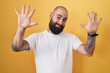 Canvas Print - Young hispanic man with beard and tattoos standing over yellow background showing and pointing up with fingers number ten while smiling confident and happy.