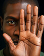 A close-up image of a young adult black man with one hand covering half of his face, revealing only one eye and creating an intimate portrait.