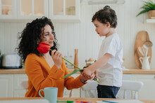 Cute Little Boy Dares To Mom Beautiful Red Tulip On Mothers Day At Kitchen. Family Moments. Children Love, Motherhood. Young Happy Woman Receiving Flower From Cute Kid Home.