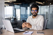 © Tetiana - Portrait of a young Indian businessman, office worker sitting at a desk and talking on the phone, smiling and looking at the camera