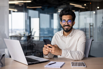 Wall Mural - Portrait of a young Indian businessman, office worker sitting at a desk and talking on the phone, smiling and looking at the camera