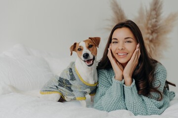 Sticker - Joyful woman in a cozy sweater shares a warm moment with her playful Jack Russell Terrier on a bed