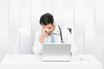 Close up portrait of tired male doctor sitting at the desktop and working on laptop in the office of modern clinic	