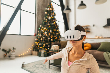 A fitness-oriented young woman practices yoga poses while wearing VR glasses by a Christmas tree.