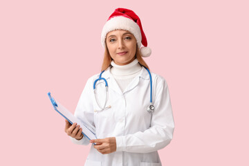 Poster - Mature female doctor in Santa hat with clipboard on pink background