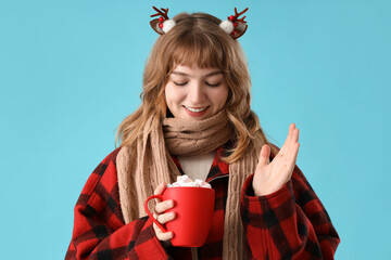 Poster - Pretty young woman in scarf holding cup of hot chocolate with marshmallows on blue background