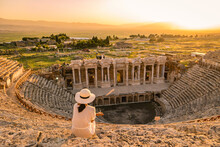 Hierapolis Ancient City Pamukkale Turkey, A Young Woman With A Hat Watching The Sunset By The Ruins Unesco Heritage. Asian Women Watching The Sunset At The Old Amphitheater In Turkey During Holiday