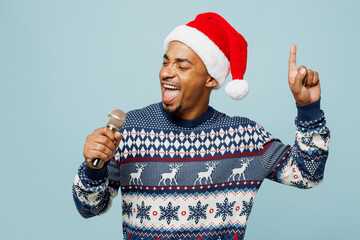 Wall Mural - Young singer man wearing knitted sweater Santa hat posing sing song in microphone at karaoke club isolated on plain pastel blue background. Happy New Year 2024 celebration Christmas holiday concept.