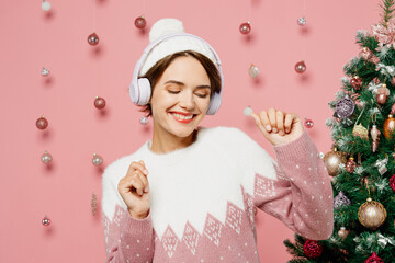 Wall Mural - Merry smiling young woman wearing white sweater hat posing listening to music in headphones dance isolated on plain pastel pink background studio. Happy New Year celebration Christmas holiday concept.