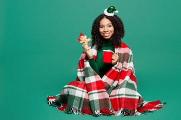 Wall Mural - Full body fun merry little kid teen girl wrapped in plaid wear hat posing drink tea eat ginger cookie isolated on plain green background studio portrait. Happy New Year celebration Christmas concept.