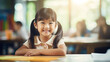 portrait of a smiling asian school girl sitting in front of school desk on a blurred classroom background with classmates