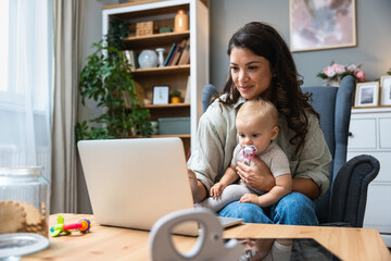 Stay at home mom working remotely on laptop while taking care of her baby. Young business mother on maternity leave trying to freelance by the desk with toddler child. Global work on computer