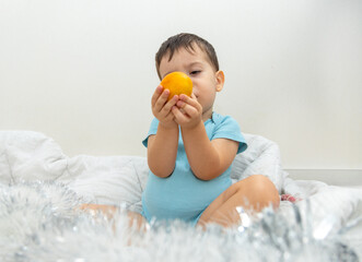 adorable baby boy on bed blanket with tangerine in hand or candy cane in heart shape. serious funny kid making grimaces,half closed eyes,cool child.merry christmas holiday at home