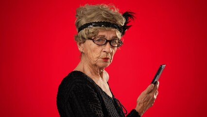 Wall Mural - Funny elderly woman walks across frame while scrolling on her smart phone, stops smiles at camera and continues across frame. Isolated on red background