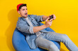 Photo of funky shocked guy wear denim jacket relaxing bean bag loosing playstation game empty space isolated yellow color background