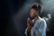 Delicate young Afro woman praying to Jesus with her eyes closed and clasped hands - profile side angle - God's rays of light shining down - Ethnic diversity and religion concept
